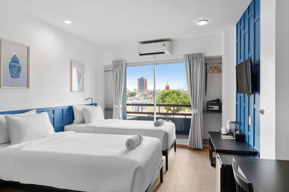 Ein Hotelzimmer mit zwei Betten, einem Balkon mit Blick auf die Stadt, einem Schreibtisch und einem an der Wand befestigten Fernseher.