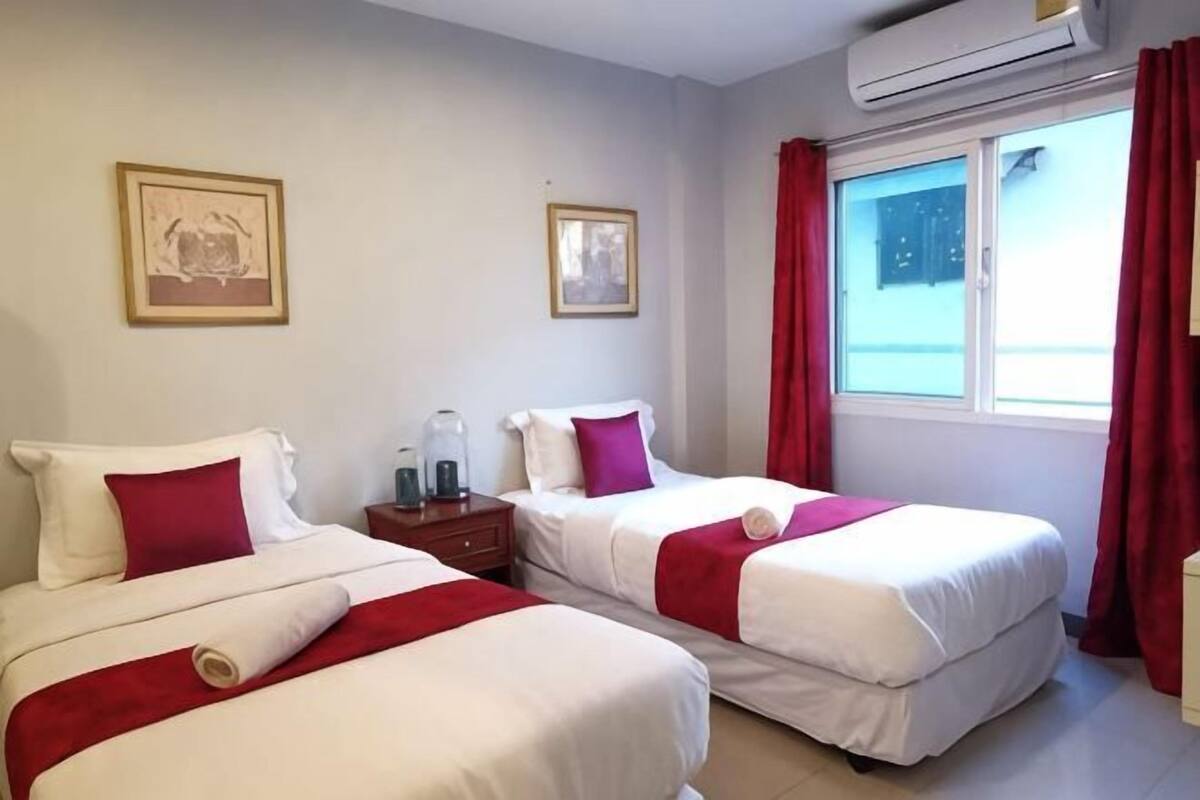 A hotel room with two beds, a window with red curtains, and an air conditioning unit.