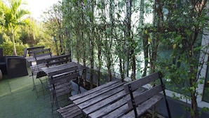 An outdoor seating area with wooden benches and tables, surrounded by lush greenery and a view of a building. - I-Beyond Residence (Bangkok)