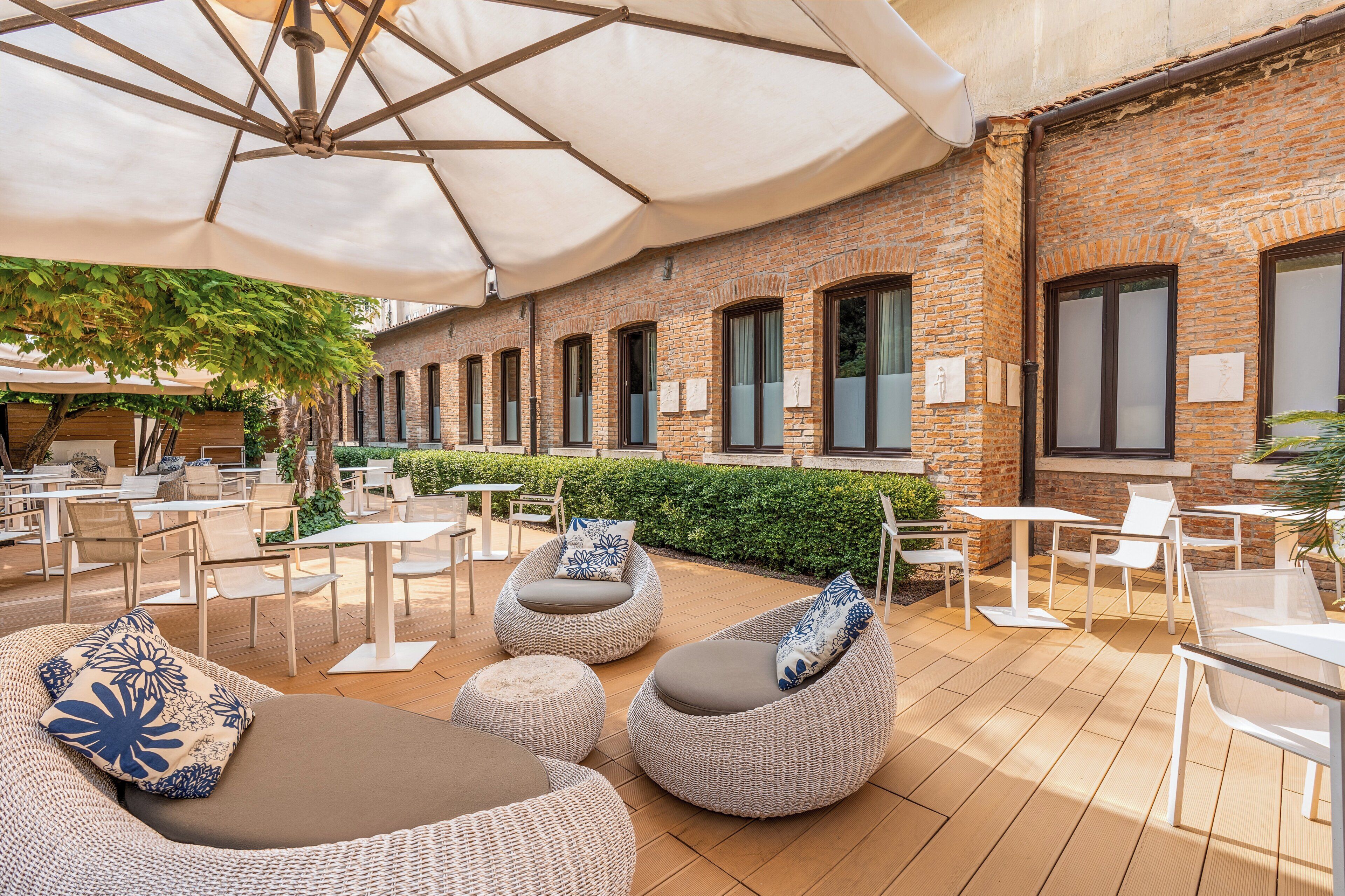 An outdoor seating area with round, cushioned chairs and tables, a brick wall, and a large umbrella providing shade.