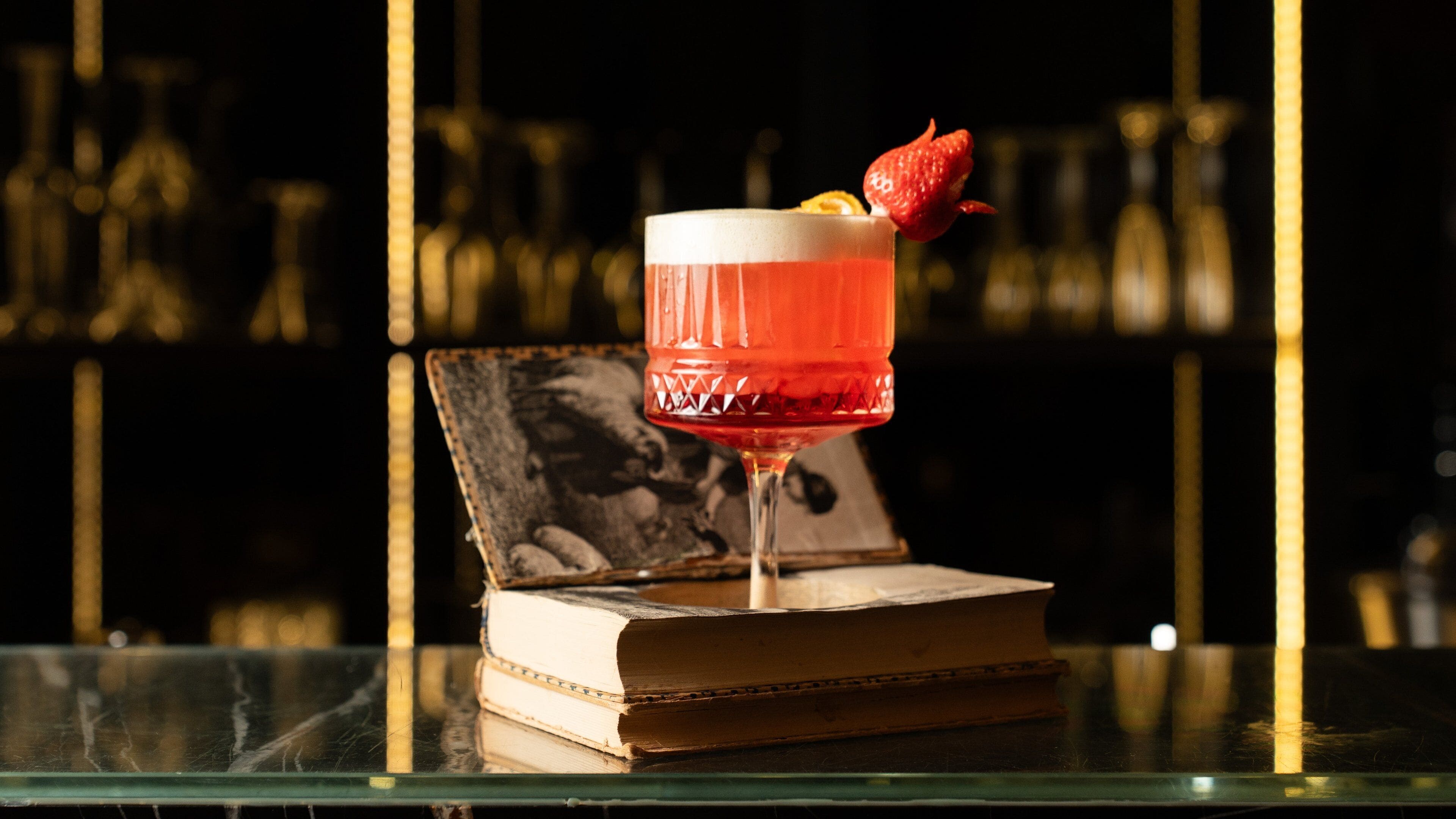 A cocktail glass with a strawberry garnish on a bar counter, a book, and a blurred background of shelves.