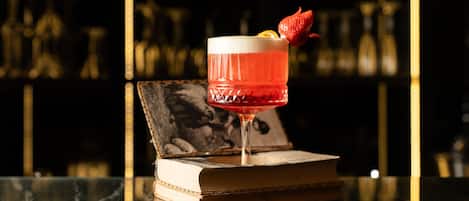A cocktail glass with a strawberry garnish on a bar counter, a book, and a blurred background of shelves.