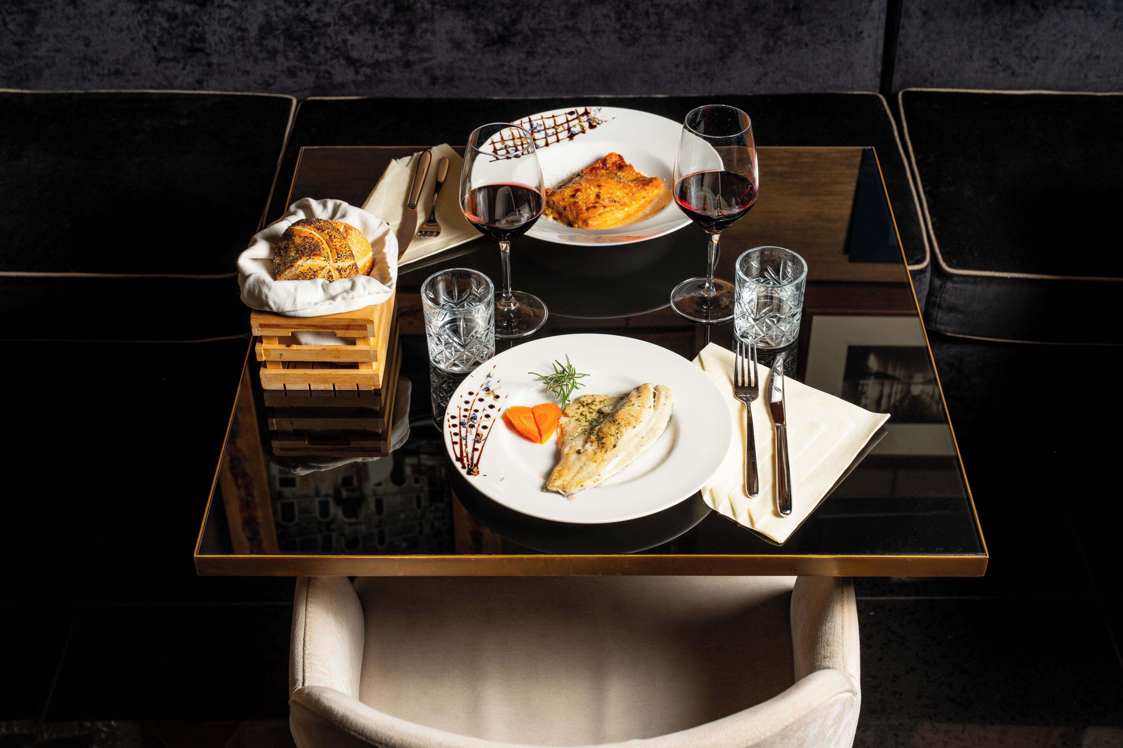 A dining table with a plate of food, two wine glasses, and a basket of bread.