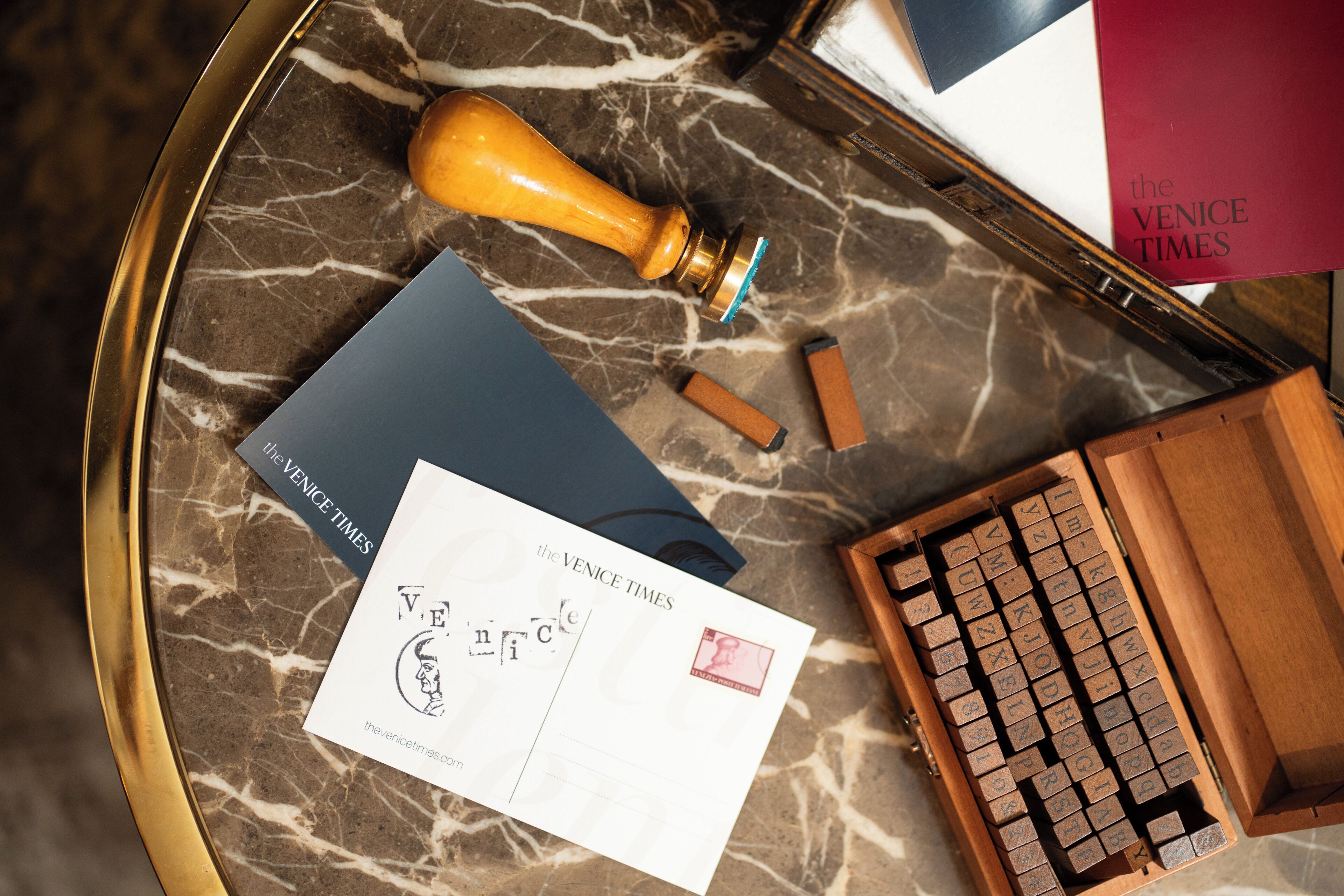 A marble surface with a wooden stamp, a postcard, a book, and a wooden box with letters.