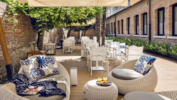 An outdoor seating area with wicker furniture and white chairs, a brick wall, and a covered section with umbrellas. - The Venice Times Hotel, Vignette Collection by IHG (Venice)