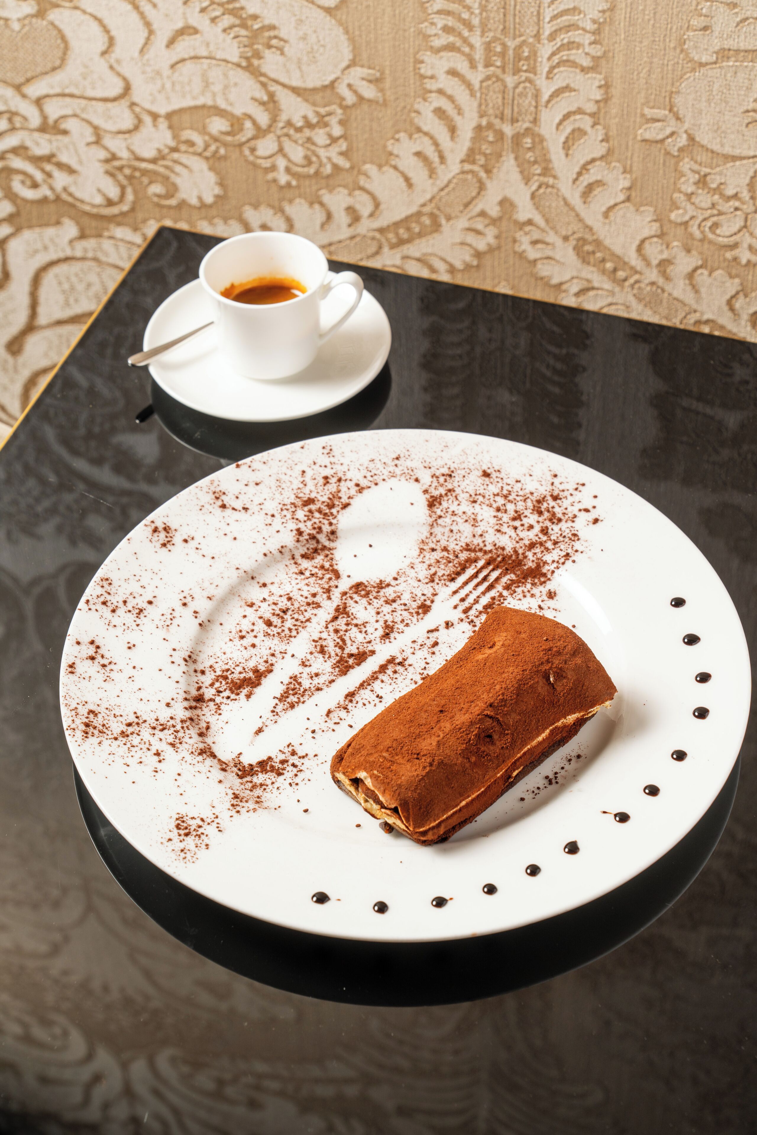 A dessert plate with a slice of tiramisu on a white plate with brown powder sprinkled on top, a cup of coffee on a saucer, and a black table with a patterned tablecloth.