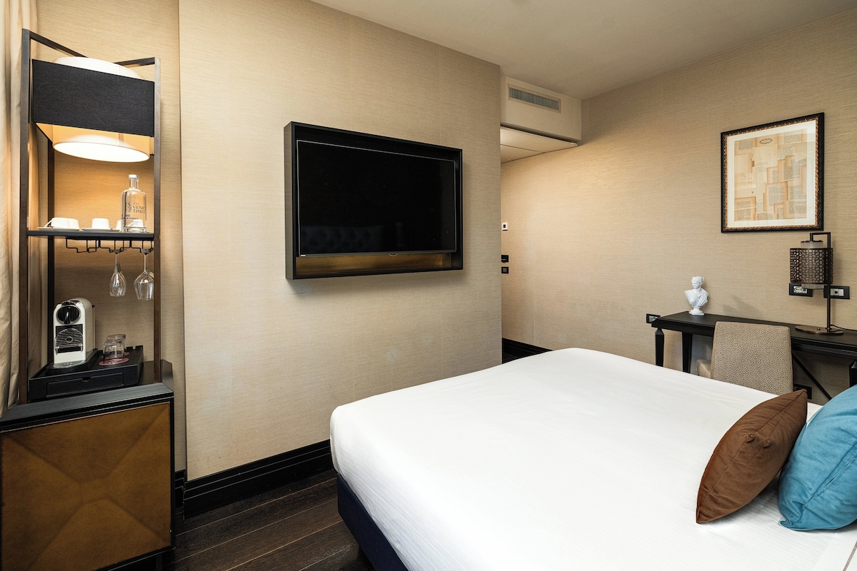 Habitación de hotel con cama, televisor de pantalla plana, escritorio con lámpara y una mesita con una botella y vasos.