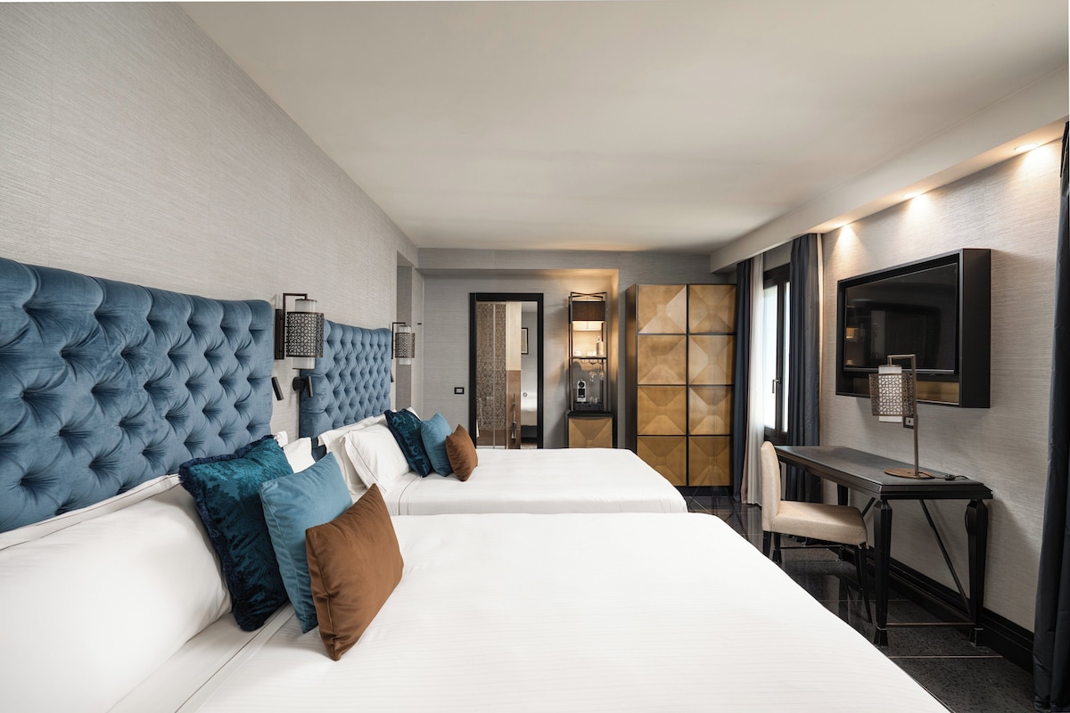 Habitación de hotel con dos camas, un escritorio, una silla, un televisor y un baño visible a través de una puerta abierta.