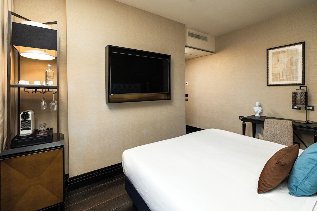 A hotel room with a bed, a flat-screen TV, a desk with a lamp, a small table with a bottle, and a framed picture on the wall.
