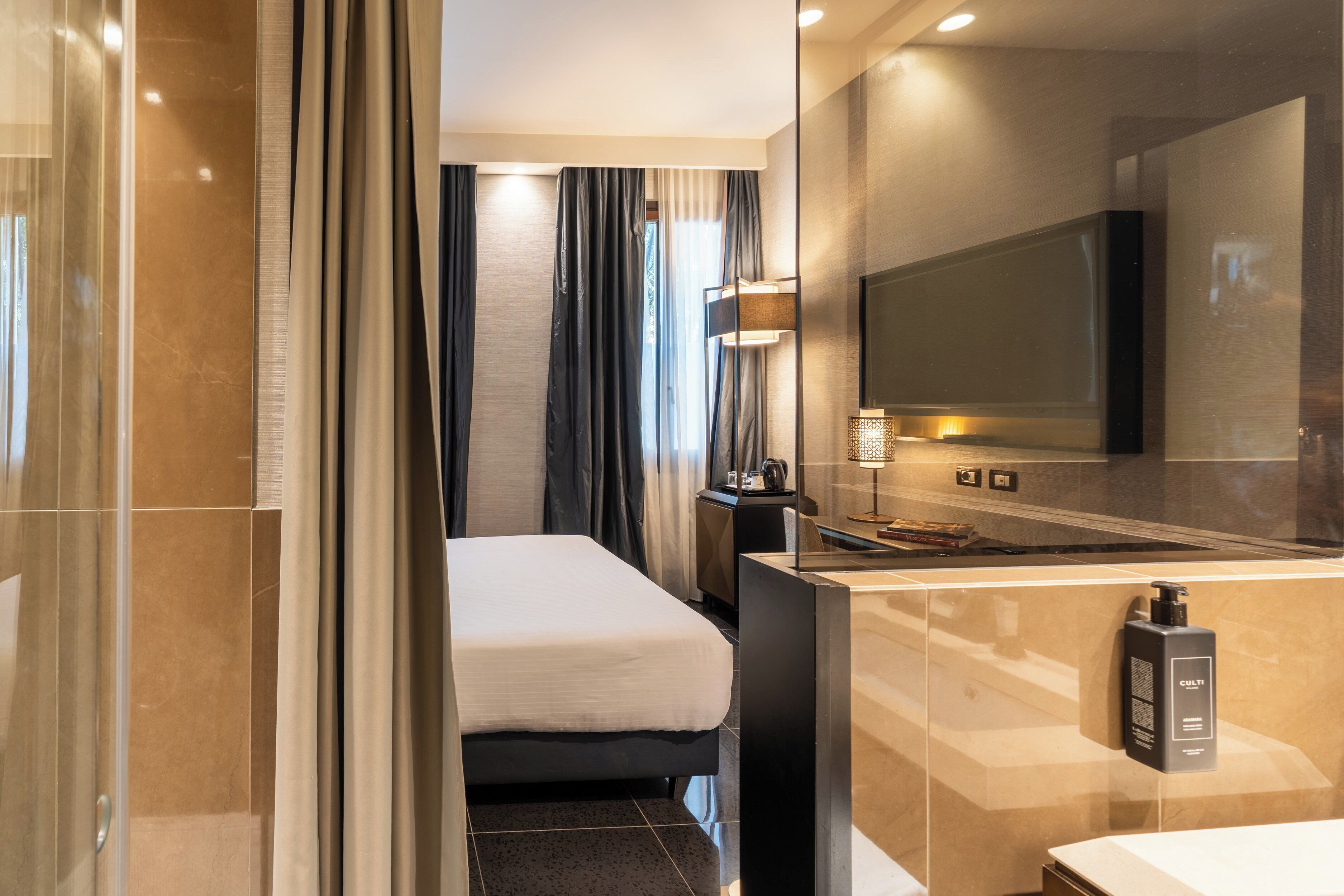 Una camera d'albergo moderna con un letto, una TV, un minibar e un bagno con uno specchio e un lavandino.