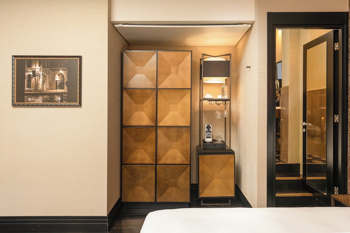 Habitación de hotel con cama, armario, un cuadro en la pared y una mesita con lámpara y botella.