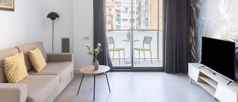 Una sala de estar moderna con un sofá, una mesita con un florero, un televisor en un mueble bajo y un balcón con sillas.