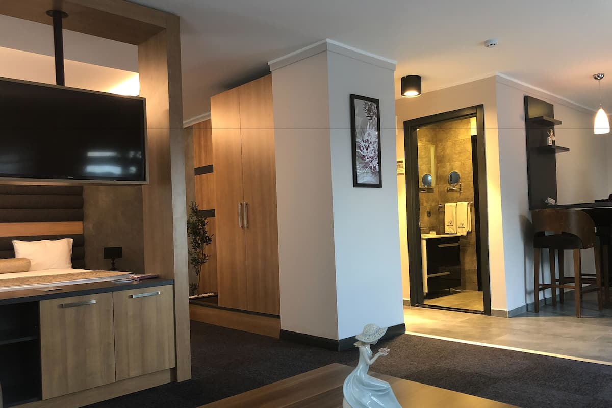 A modern hotel room with a bed, wooden wardrobe, a small table with a decorative vase, a TV mounted above the bed, and a kitchenette area.