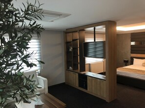A modern bedroom with a built-in TV, a bedside table, and a large window with blinds. - Kalevera Hotel (Edirne)