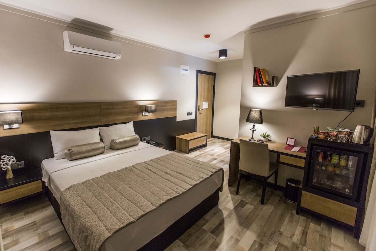 A hotel room with a bed, bedside tables, a desk, a TV, and a minibar.