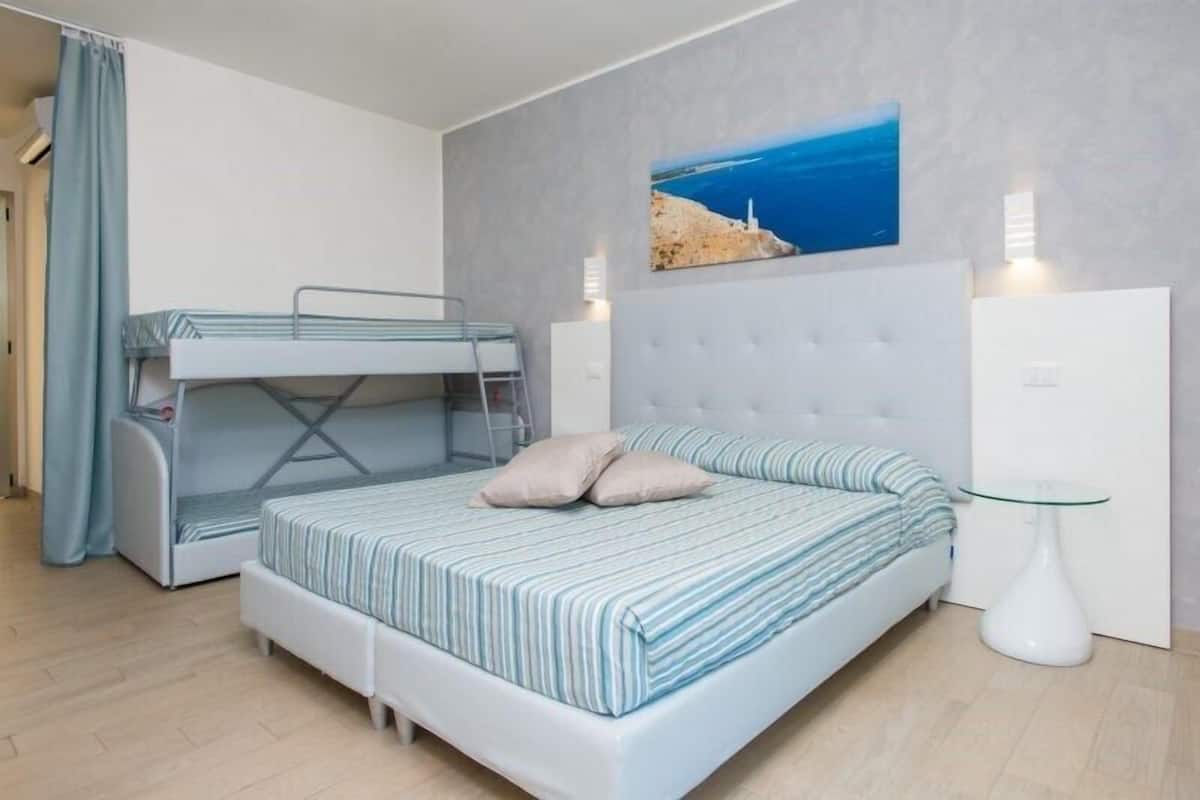 A bedroom with a bunk bed, a single bed, a glass table, and a painting of a coastal scene.