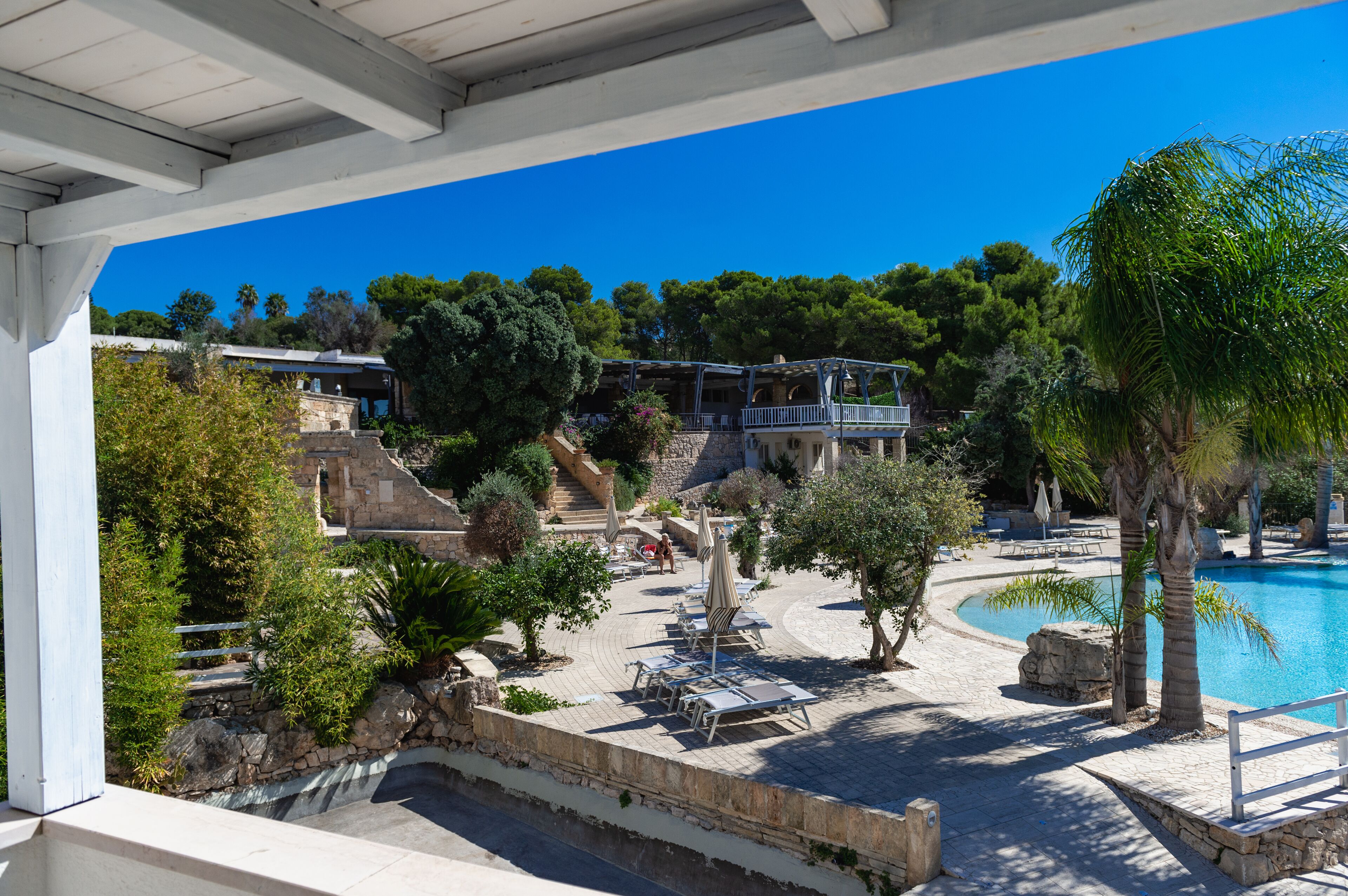 A poolside area with lounge chairs and umbrellas, a stone pathway, and a view of a building with a balcony overlooking the pool.