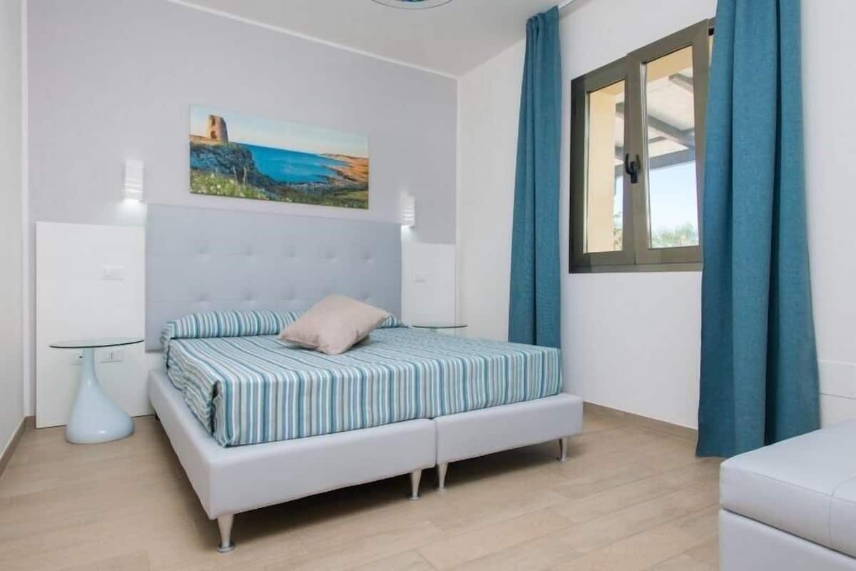 A bedroom with a bed, a painting, a window with curtains, and a bedside table.