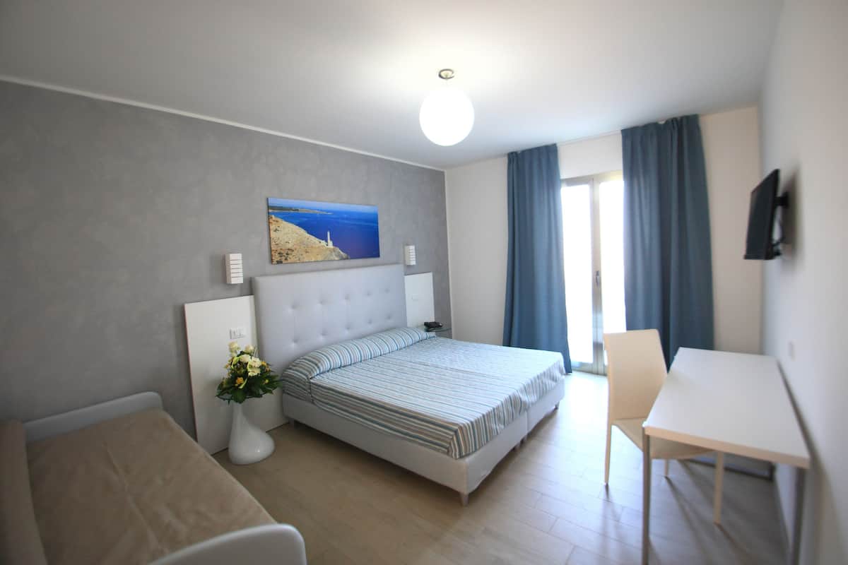 A modern hotel room with a bed, bedside table, chair, and a desk.