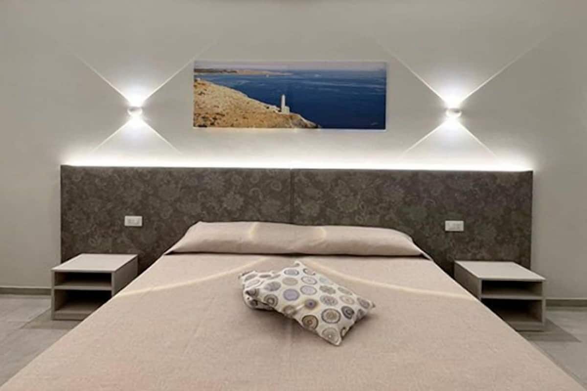 A modern bedroom with a large bed, a headboard with built-in lighting, and a framed picture of a coastal landscape.