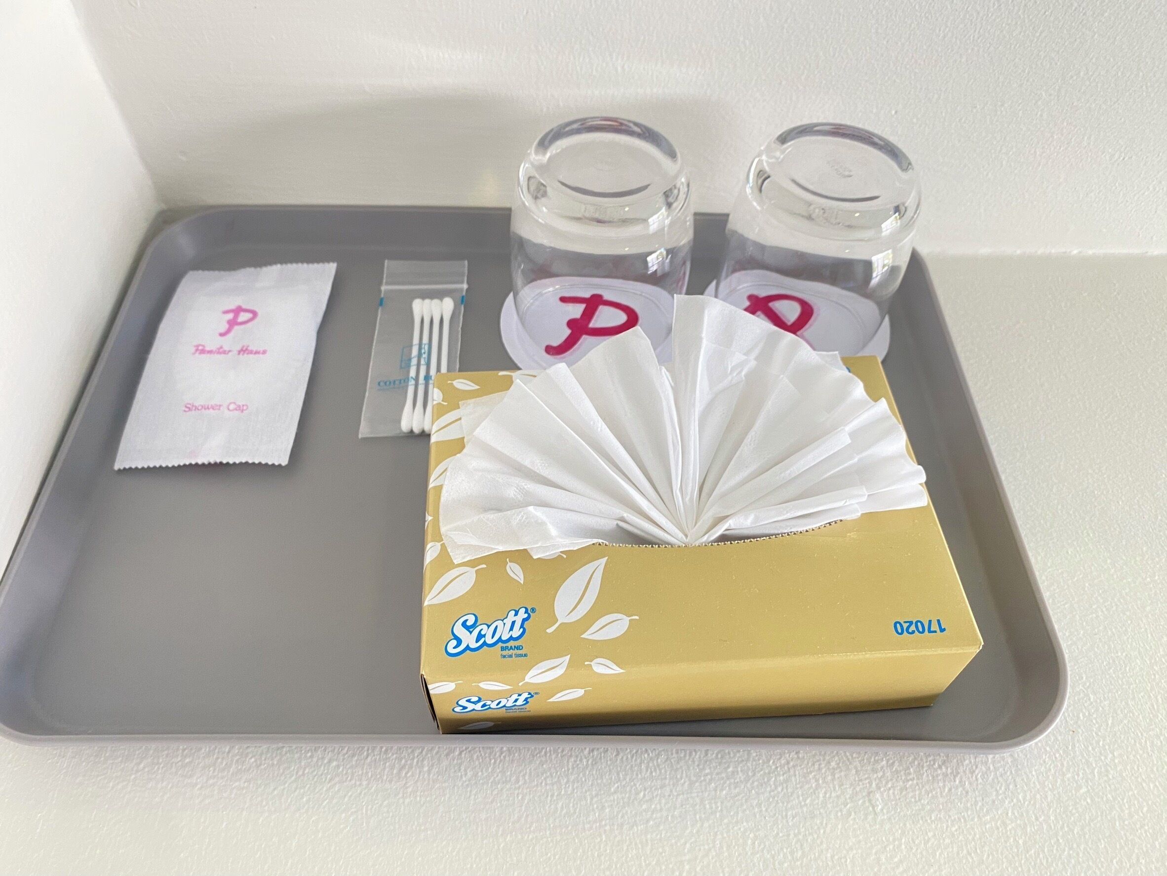 A tray with a box of Scott brand facial tissue, cotton swabs, a shower cap, and two glass containers with a pink 'P' logo.