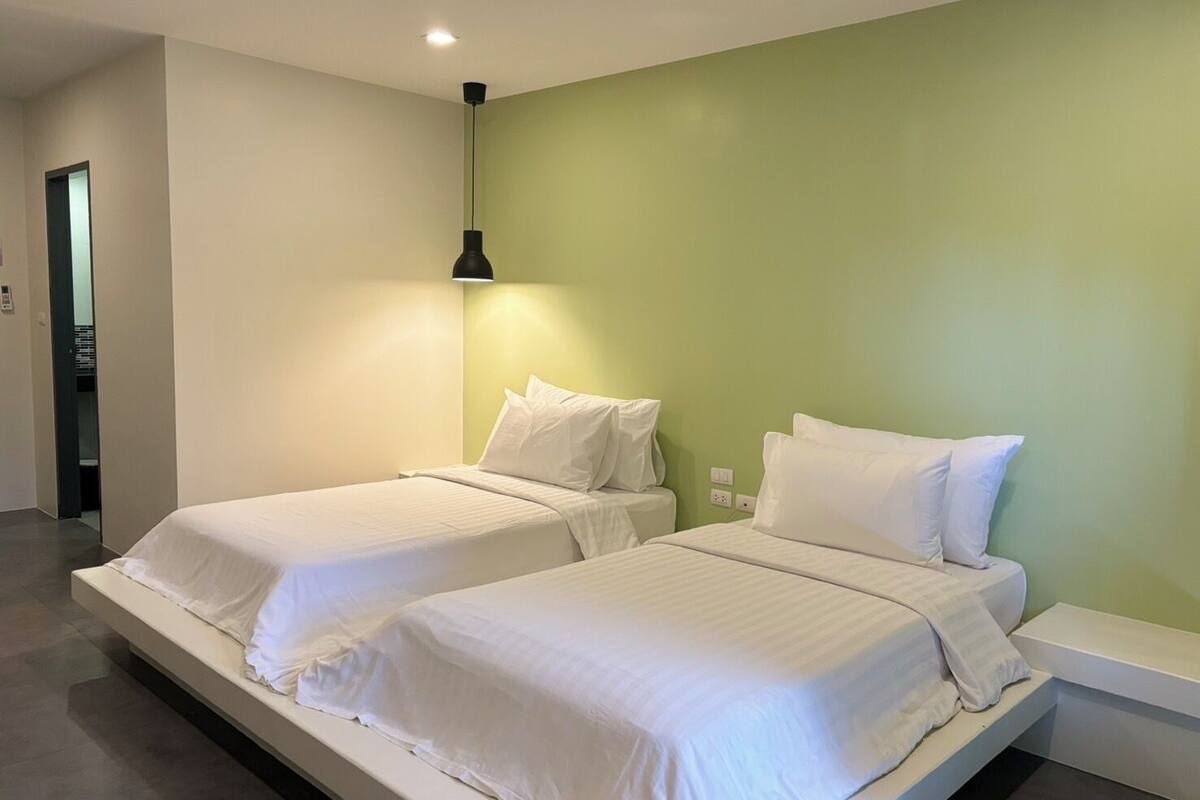 Una habitación de hotel moderna con dos camas individuales, una pared de acento verde y una lámpara colgante sobre las camas.