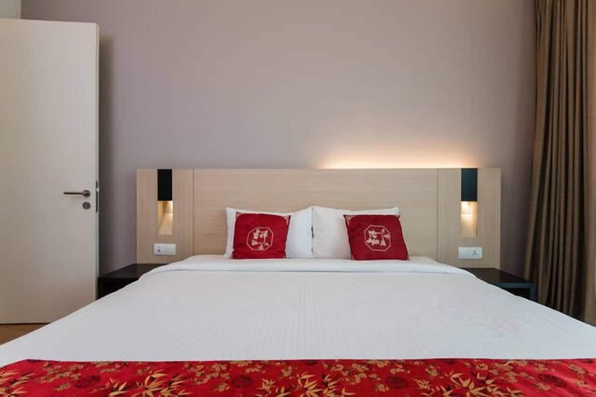 A hotel room with a bed, two red pillows, a white bedspread, a red floral blanket, a wooden headboard, bedside lamps, and a door.