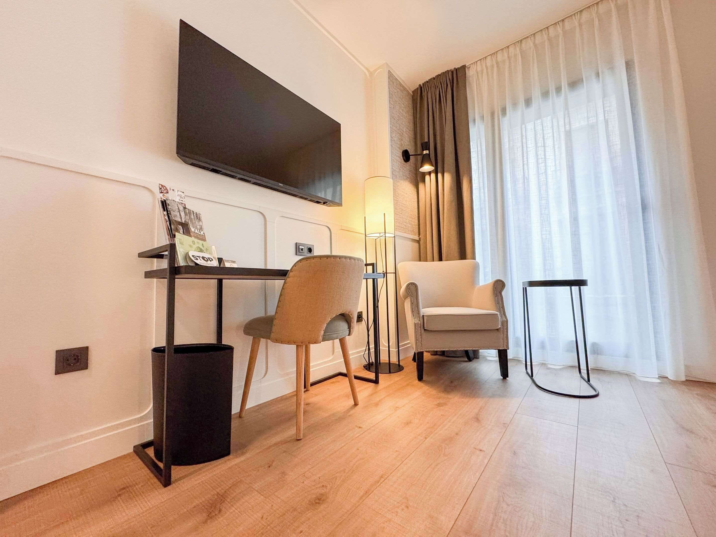 Habitación de hotel moderna con televisor de pantalla plana montado en la pared, un escritorio con una silla, una lámpara de pie y un gran ventanal con cortinas.