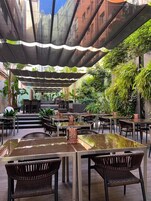 Un área de comedor al aire libre con mesas y sillas, rodeada de follaje y un diseño arquitectónico moderno.