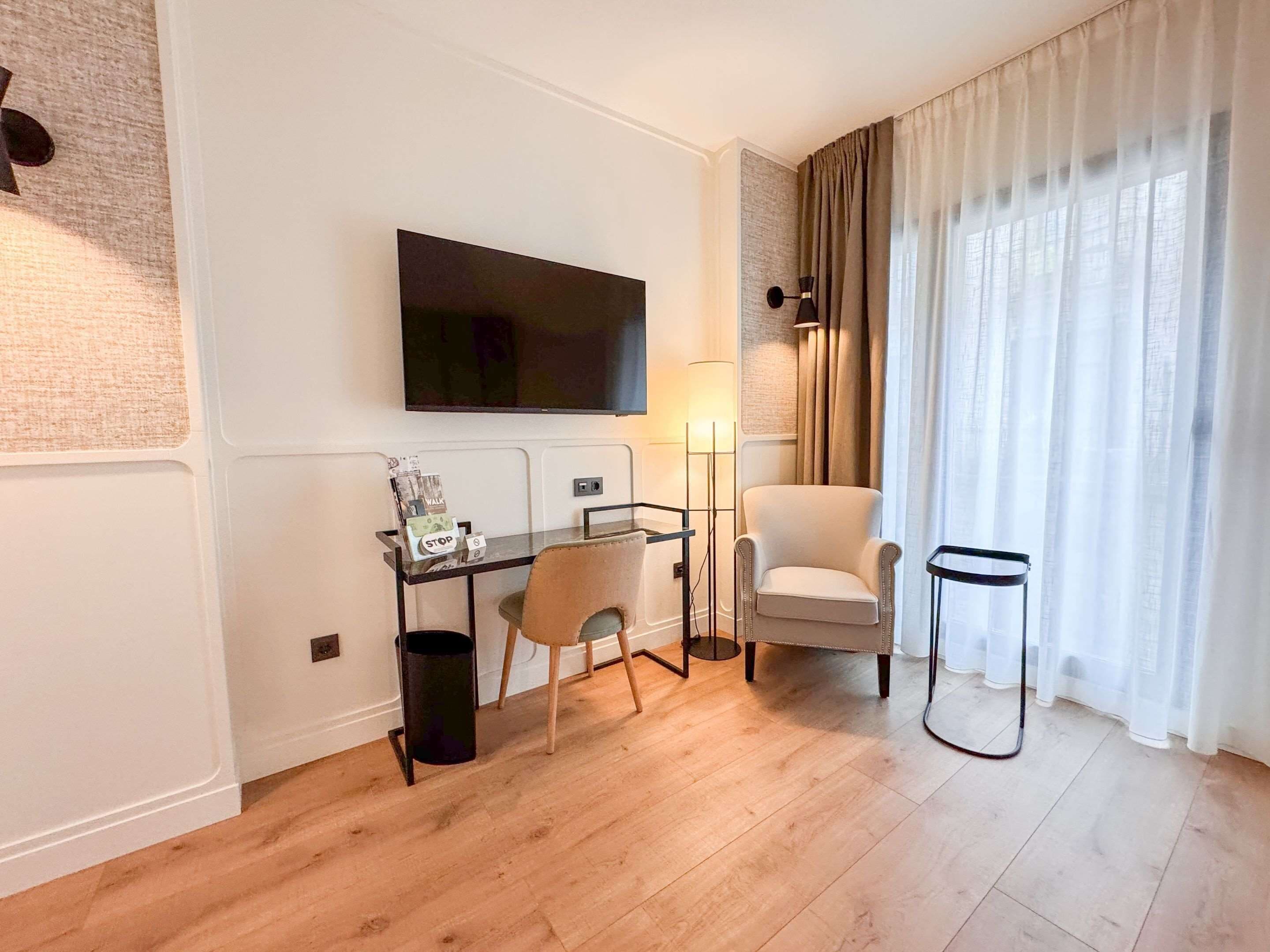 Una habitación de hotel moderna con televisión de pantalla plana montada en la pared, un escritorio con silla, una mesita auxiliar pequeña y un ventanal con cortinas traslúcidas.
