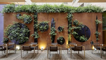 Un área de comedor al aire libre con una terraza de madera, sillas y mesas de metal. Hay macetas circulares con follaje en la pared detrás de la zona de estar.
