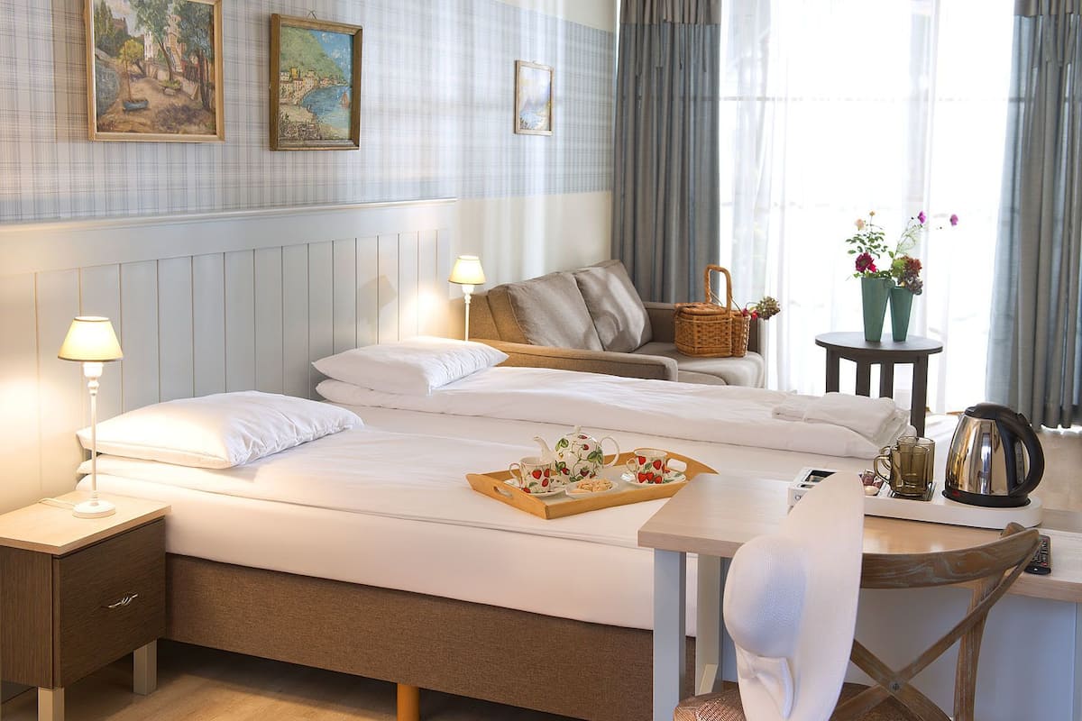 A hotel room with a bed, bedside tables, a desk, a chair, a lamp, a tray with breakfast items, a kettle, and a window with curtains.