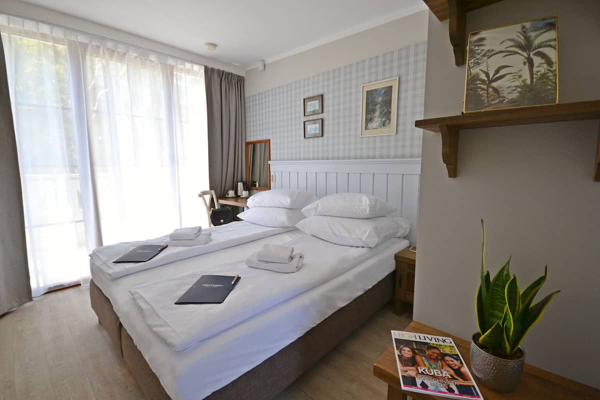 A hotel room with a large bed, a bedside table with a magazine, a potted plant, and a window with curtains.