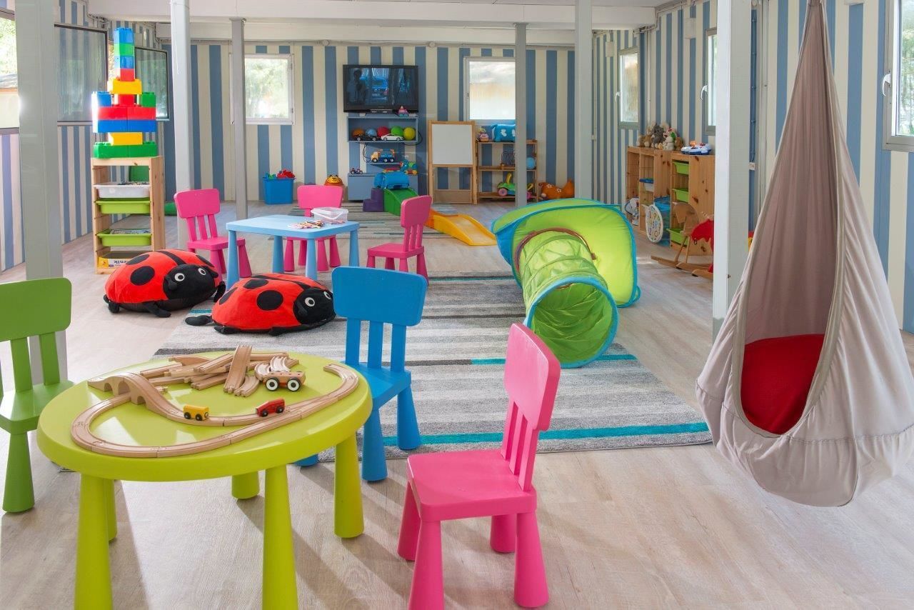 Ein helles Kinderzimmer mit Spieltisch, Stühlen, einer Hängematte, einer Rutsche und einer Spielanlage.