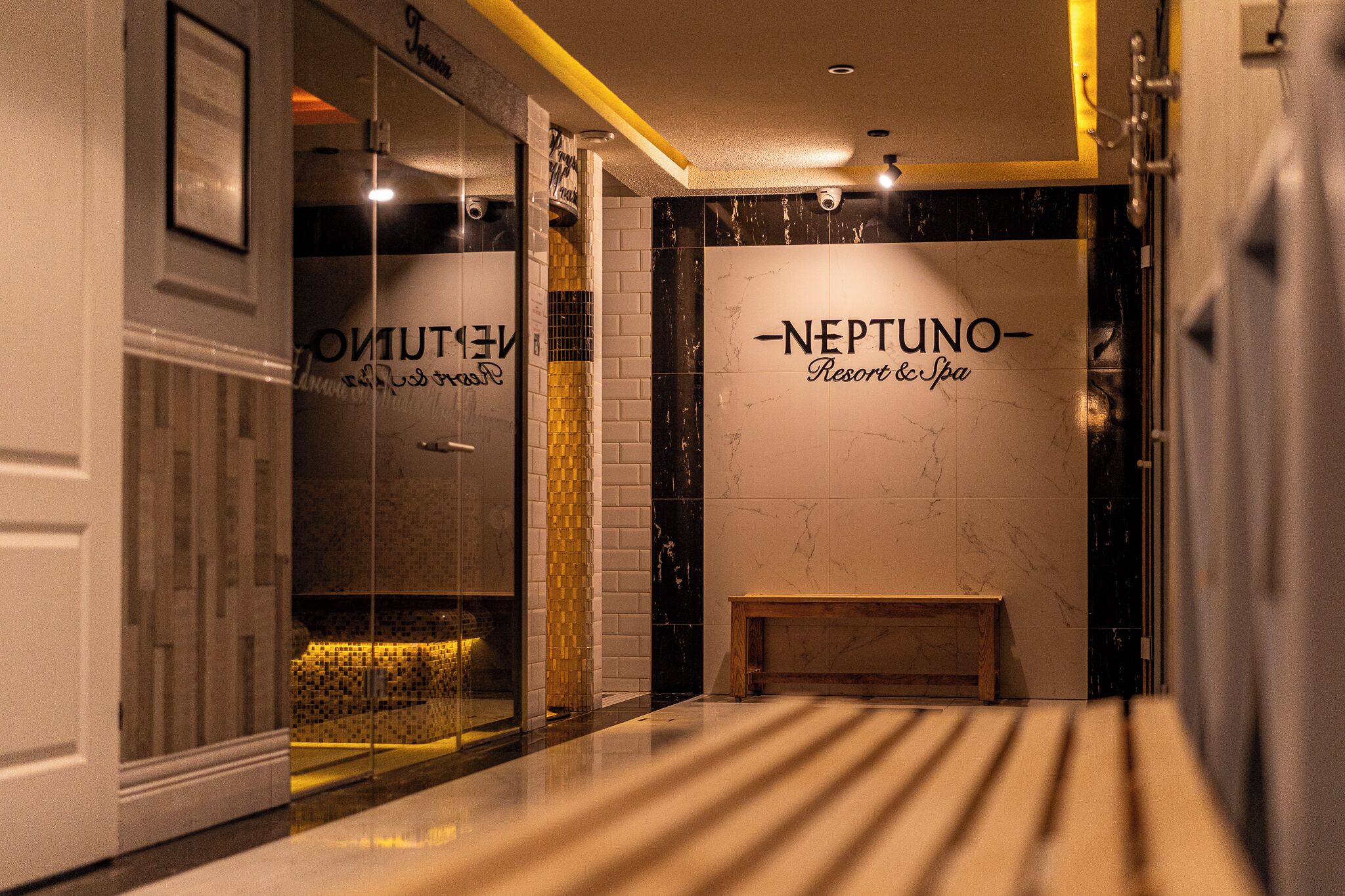 Un pasillo con un banco, un cartel que dice 'NEPTUNO Resort & Spa' y una pared de vidrio con reflejos.