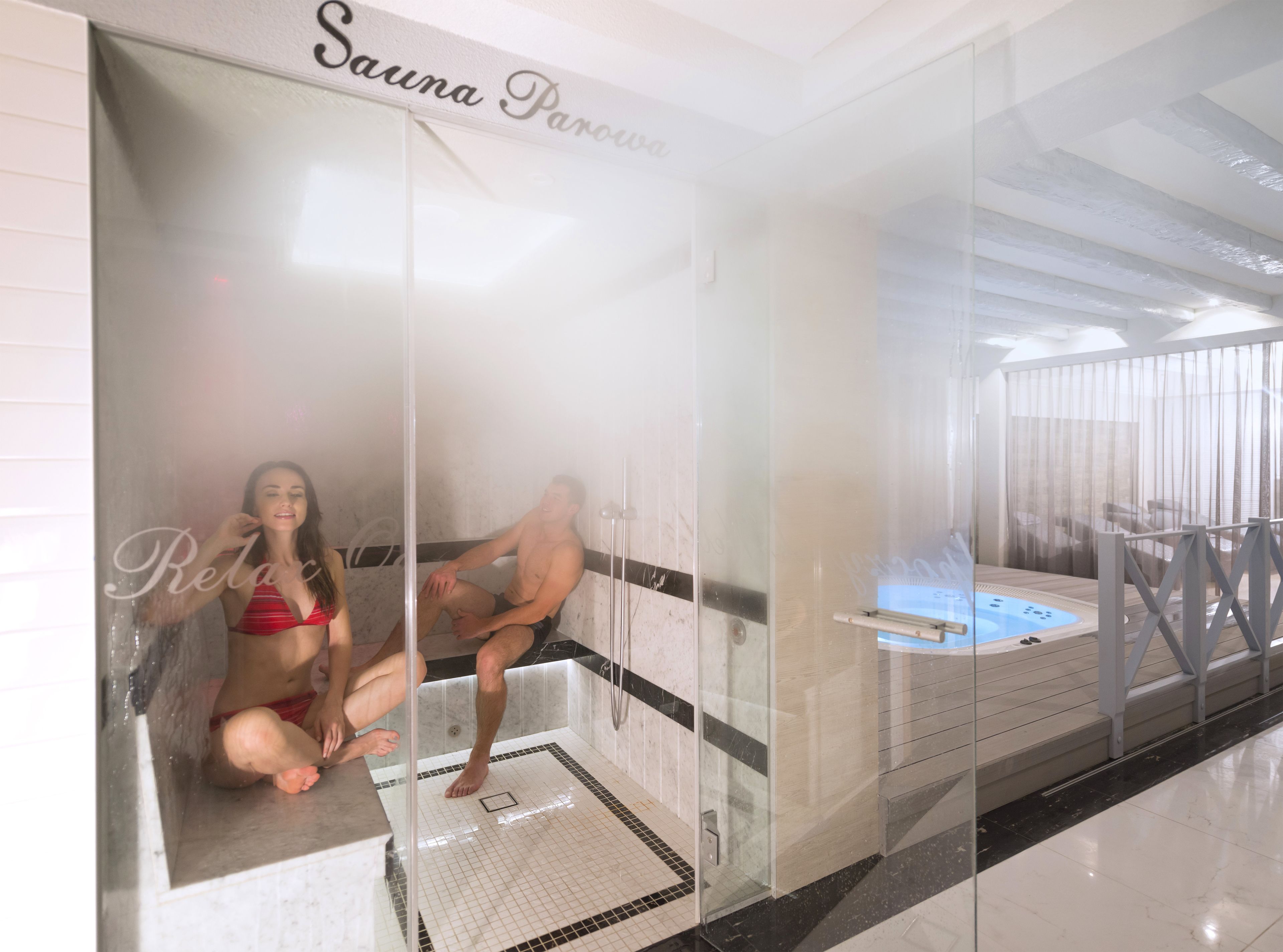 Zona de sauna con paredes de vidrio, piso de baldosas y una bañera de hidromasaje visible.