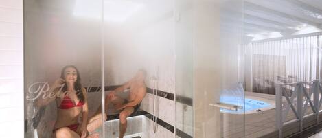 Zona de sauna con paredes de vidrio, piso de baldosas y una bañera de hidromasaje visible.