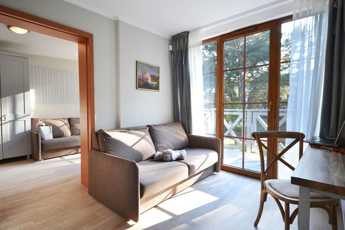 A modern living room with a sofa, a dining table, and a chair. There is a large window with curtains, a painting on the wall, and a view of trees outside.