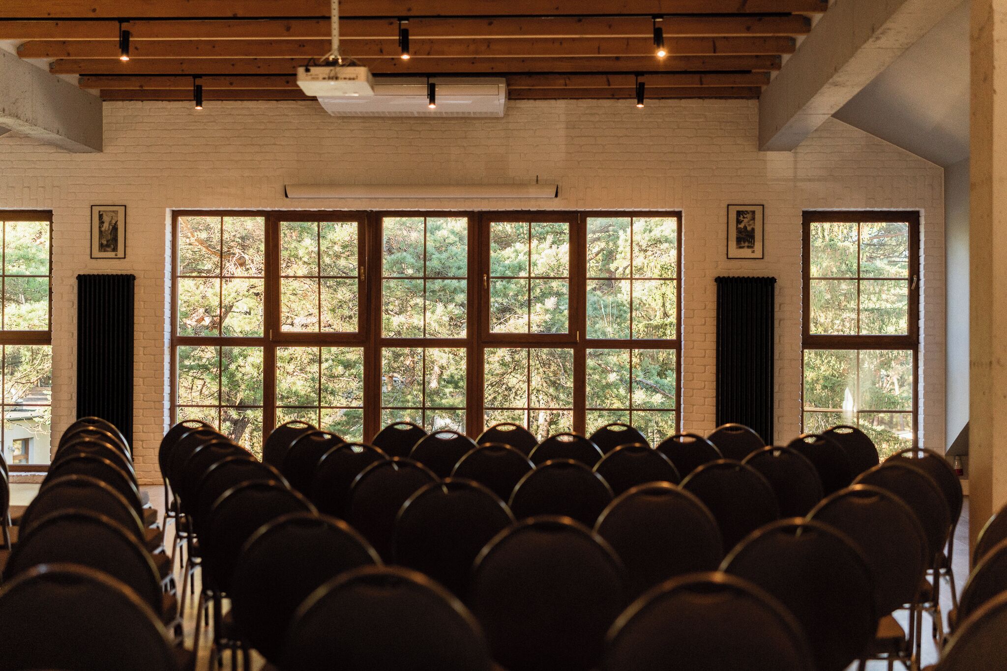 Un salón de conferencias con filas de sillas, ventanas amplias que dan a vistas de árboles y una pared de ladrillo.