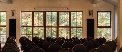 A conference room with rows of chairs, large windows with a view of trees, and a brick wall.