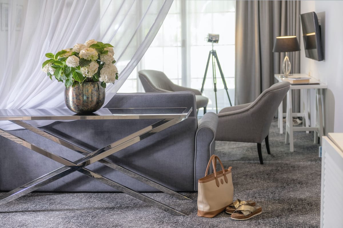 A modern hotel room with a grey sofa, a metal-framed table, a vase of white flowers, a handbag, and a pair of sandals on the floor.