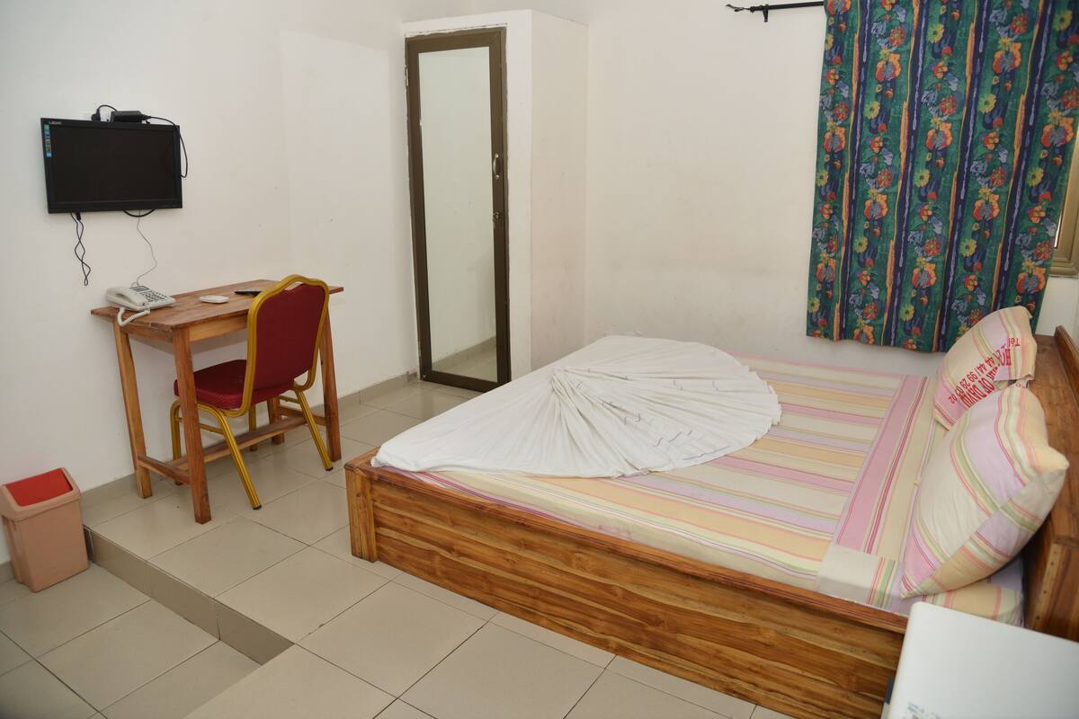 A bedroom with a wooden bed, a desk with a chair, a television mounted on the wall, and a door.