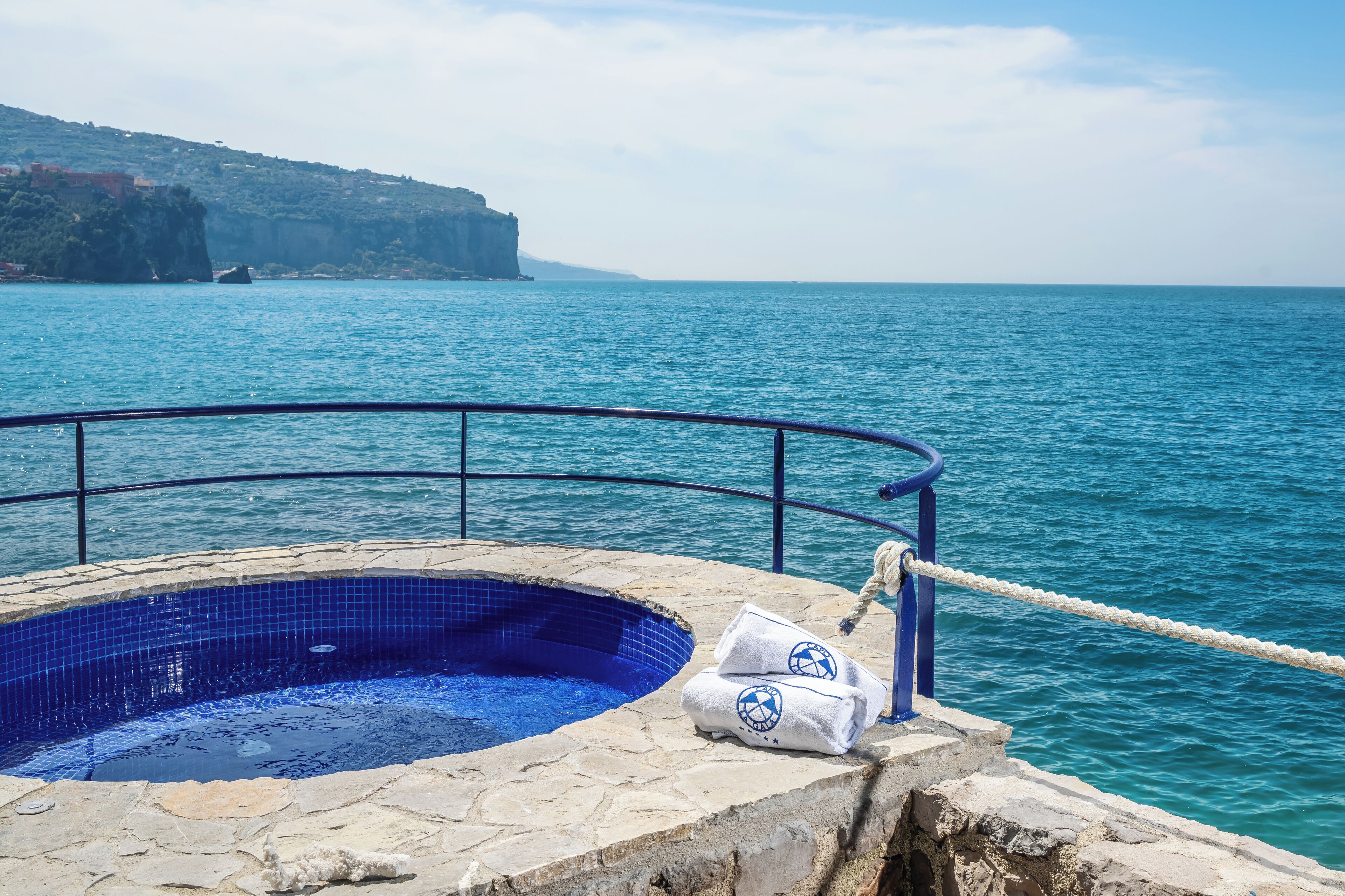 Una piscina de piedra con interior de azulejos azules, rodeada por una terraza de piedra y una barandilla metálica, con vistas a una gran extensión de agua y una costa rocosa en la distancia.