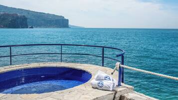 Una piscina de piedra con interior de azulejos azules, rodeada por una terraza de piedra y una barandilla metálica, con vistas a una gran extensión de agua y una costa rocosa en la distancia.