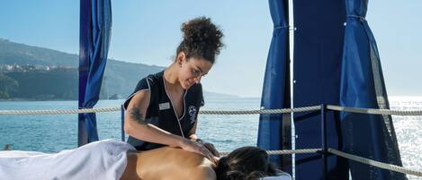 A massage therapy session on a beachside platform with a rope barrier and a view of the sea and hills in the background.