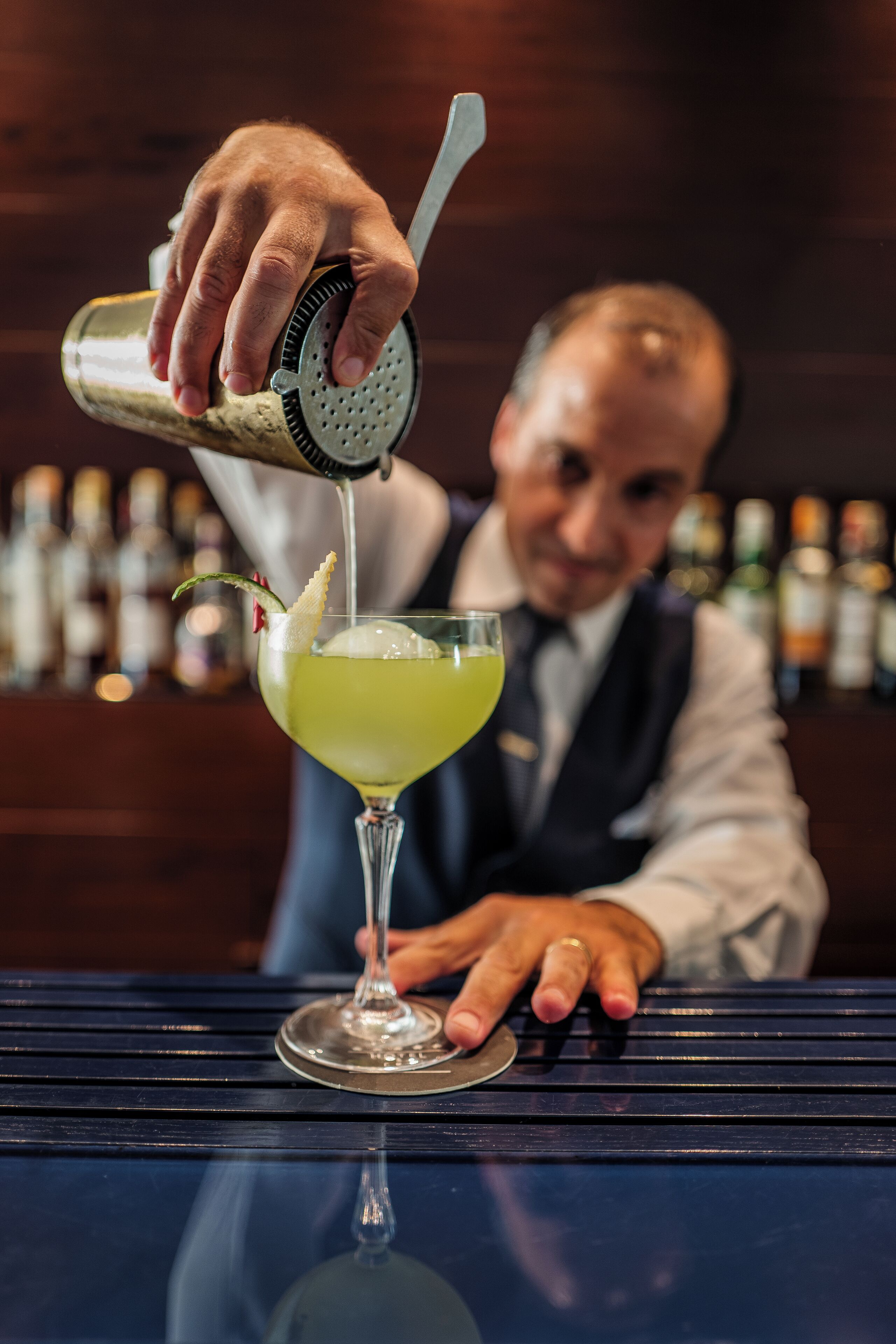 A bartender pouring a drink into a martini glass with a lemon twist garnish.