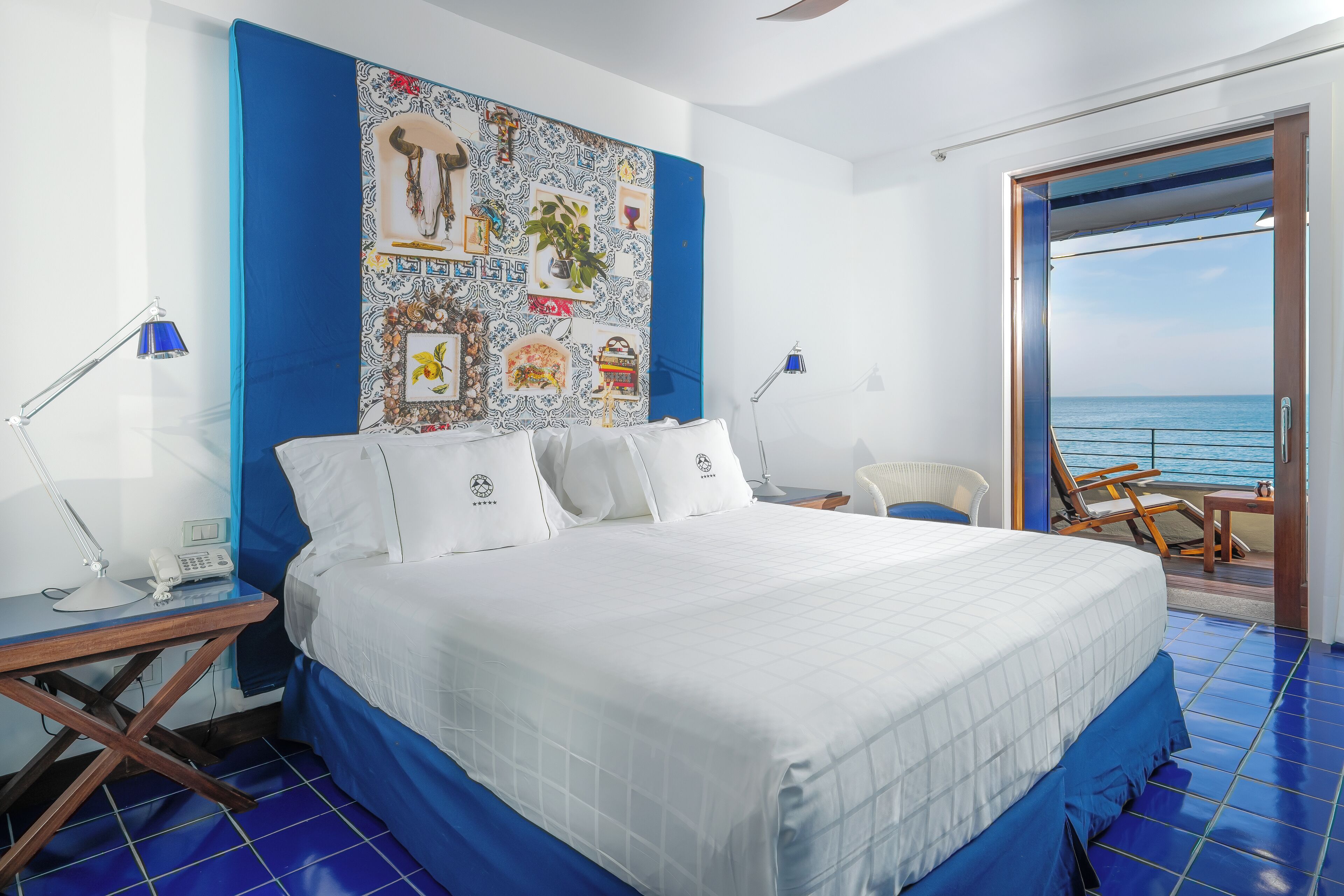 A hotel room with a large bed, a blue wall with a decorative tapestry, a balcony with a view of the sea, and a desk with a lamp and phone.