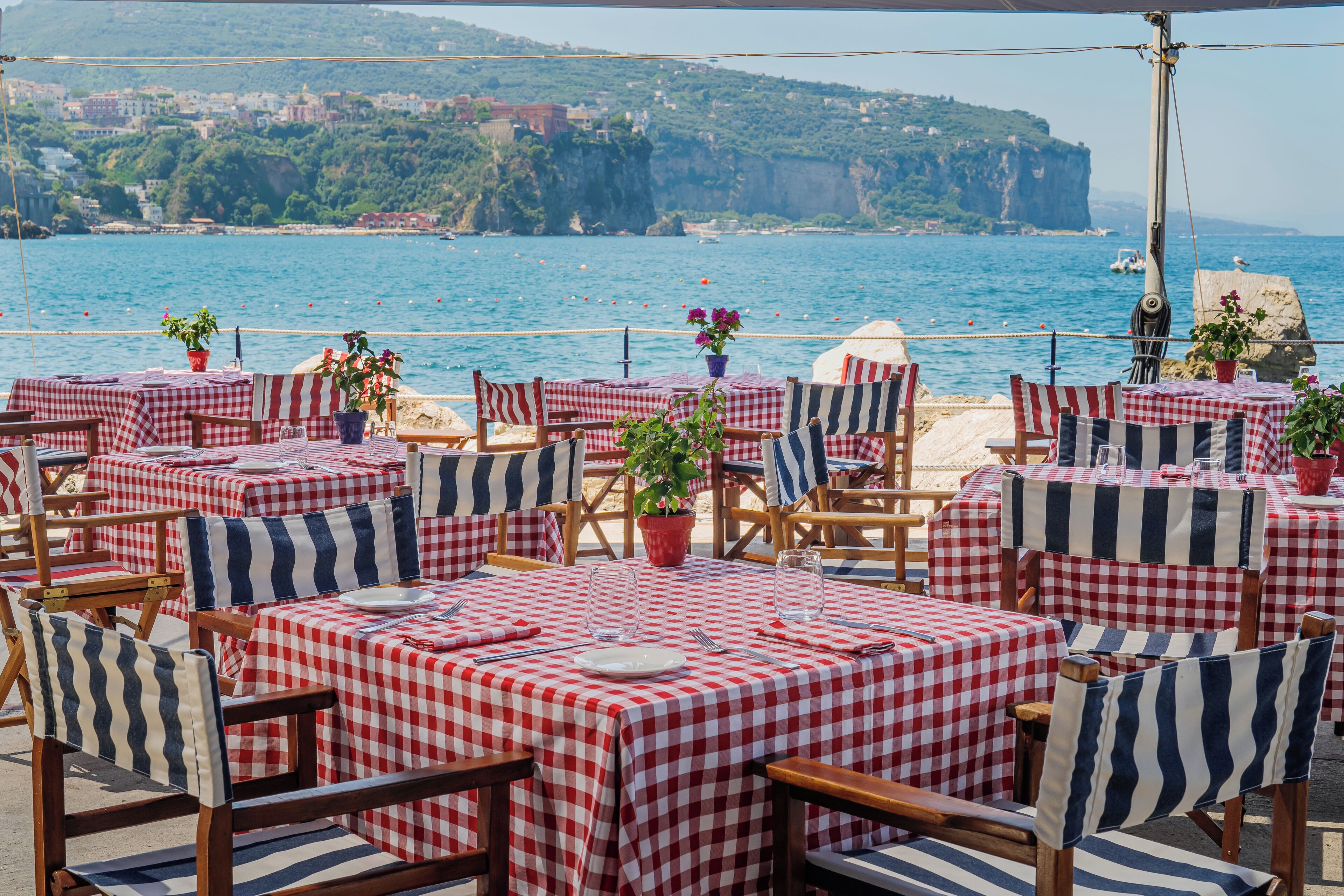 A seaside restaurant with checkered tables and striped chairs, overlooking the ocean and a cliff.