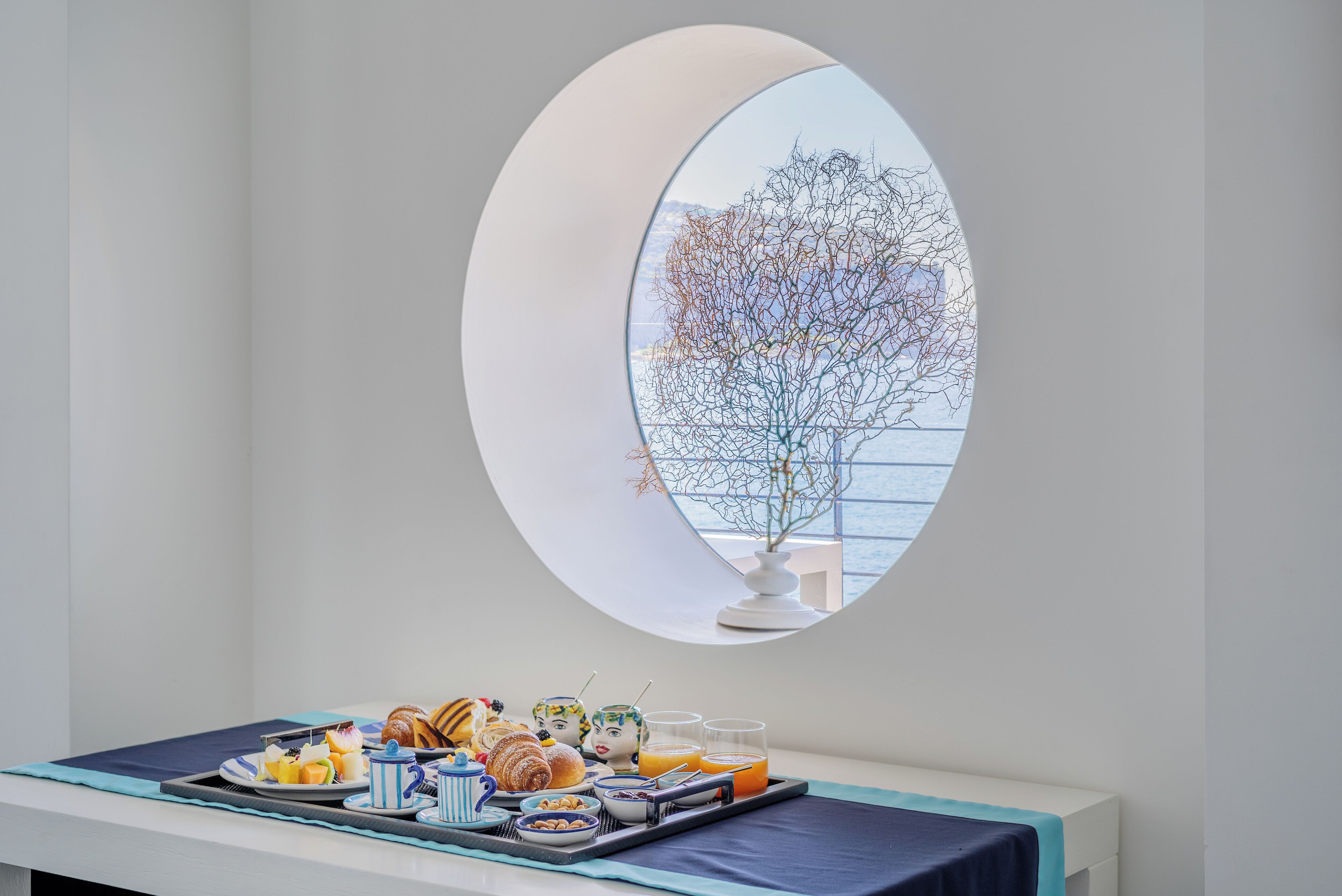 A breakfast setup with pastries, fruits, and juice on a tray, viewed through a round mirror reflecting a tree and a balcony.