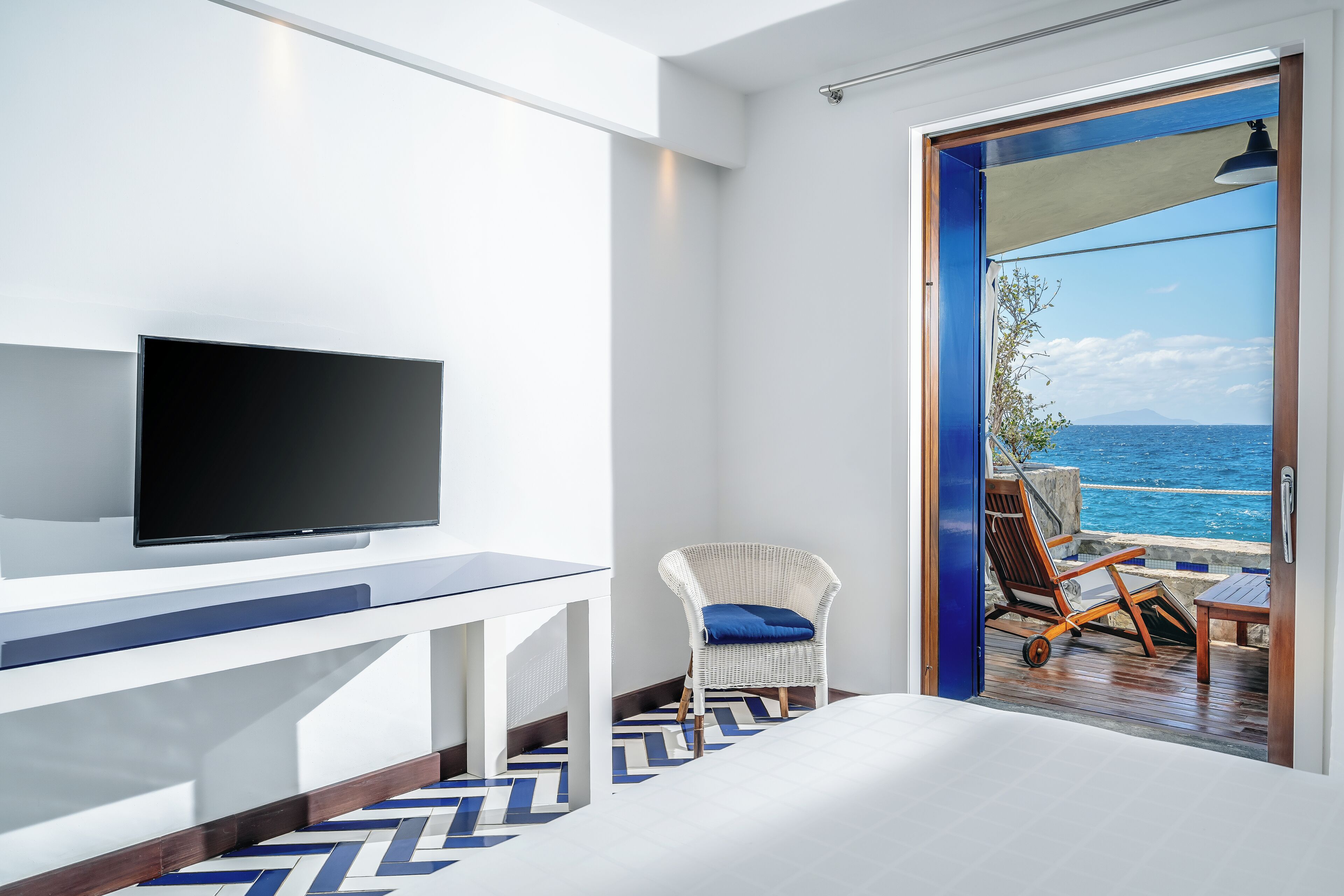 Une chambre d’hôtel moderne dotée d’une grande télévision, d’une chaise blanche avec un coussin bleu et offrant une vue sur l’océan par une porte vitrée.